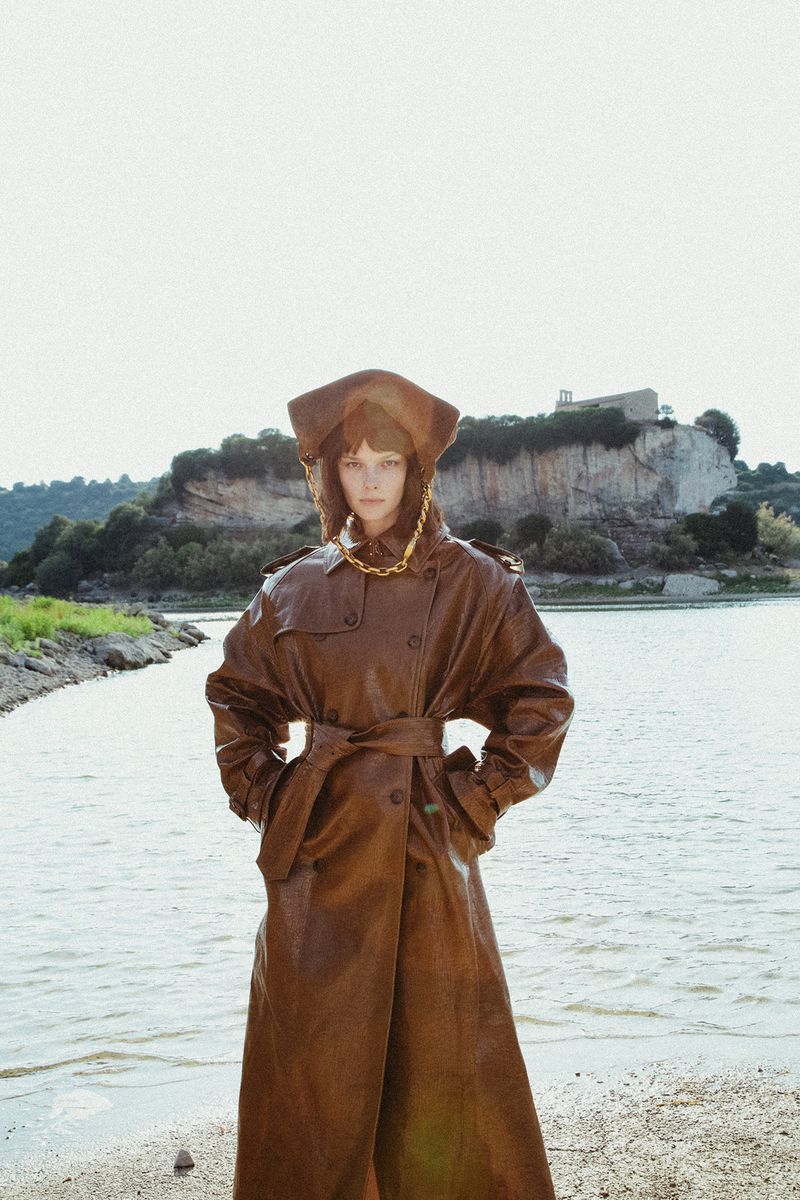 Model wearing brown leather coat and matching head covering standing outdoors near a lake and rocky cliff for Flair Germany