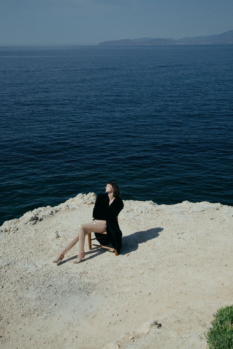 Adult female in a black coat sitting on a wooden chair on a white stone cliff overlooking a deep blue ocean on a sunny day.