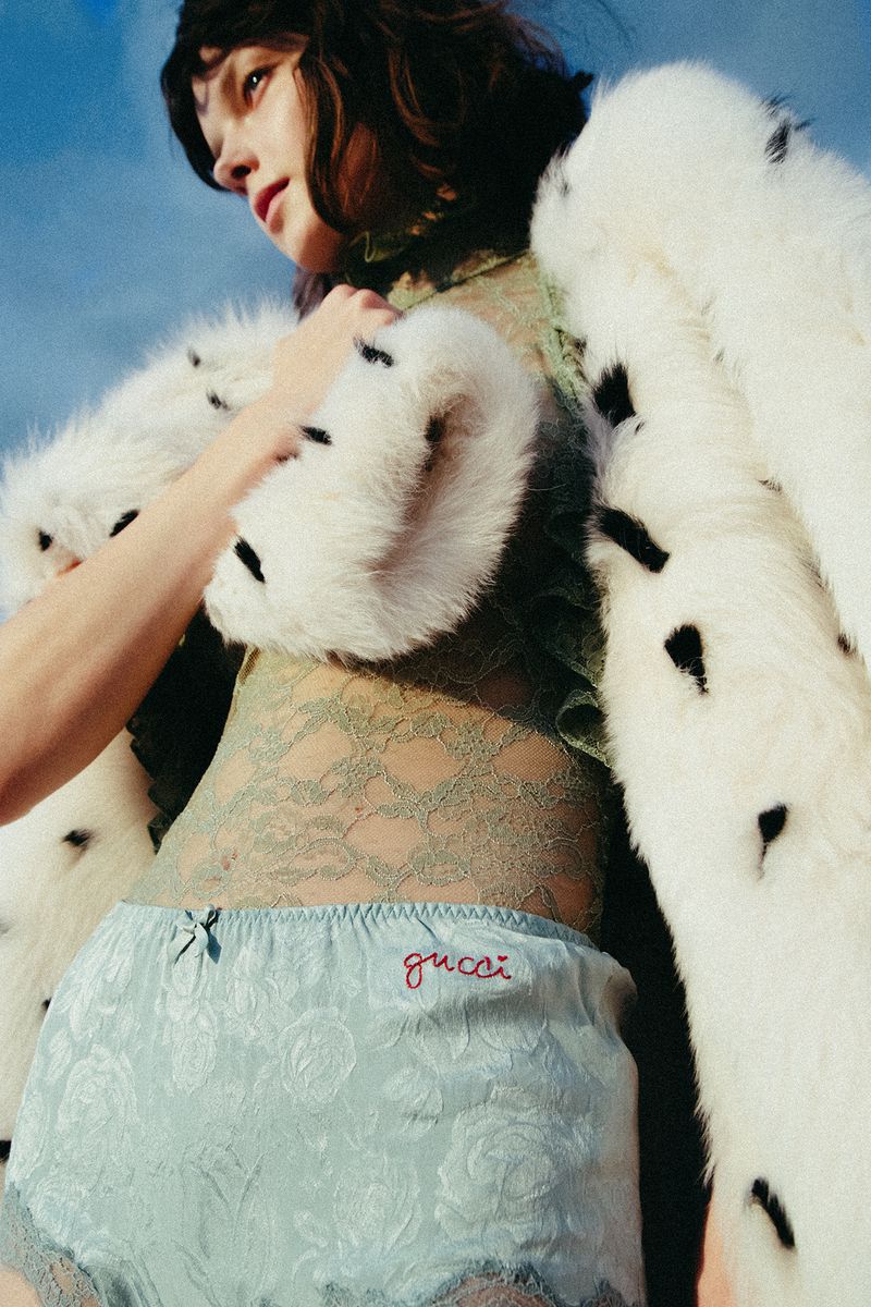 A woman in light-blue shorts with 'Gucci' embroidered, paired with a lace top and a white fur coat, stands against a soft blue sky.