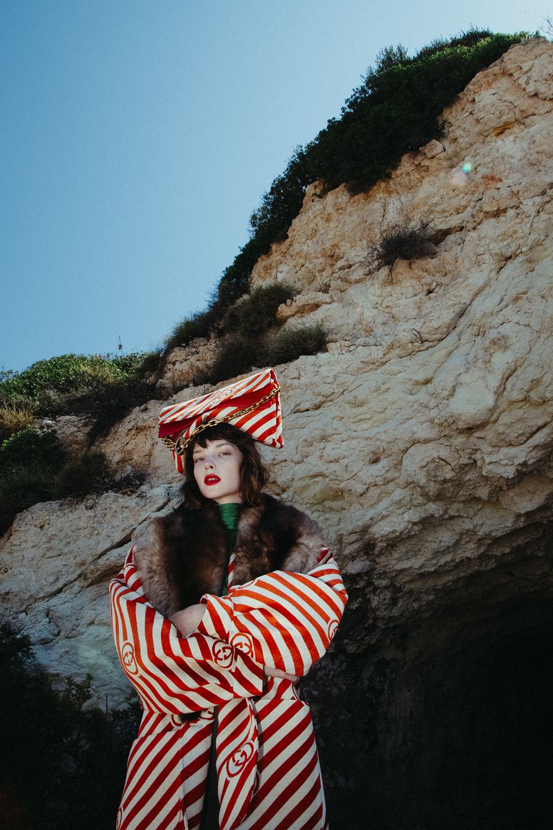 A woman poses outdoors in a red and white striped coat and matching hat against a rocky cliff under a clear blue sky in daylight.