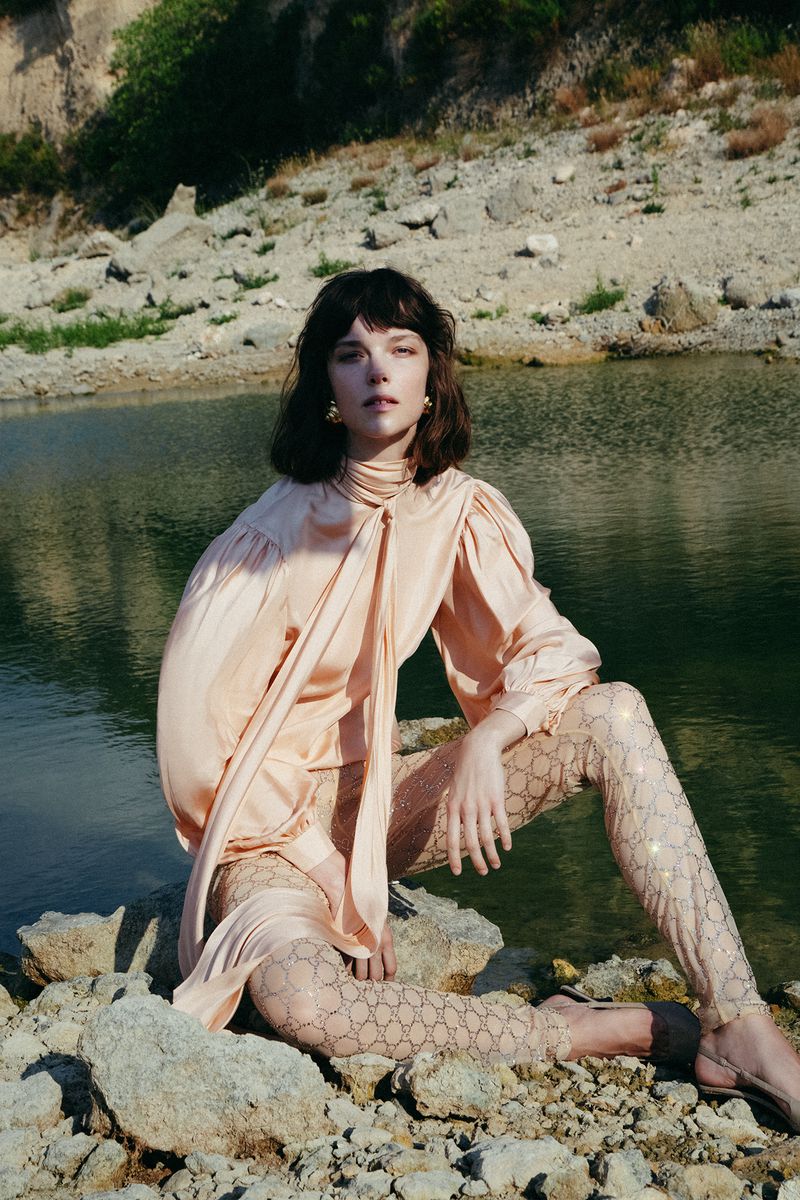 A woman with dark hair poses in a peach top and patterned leggings on rocks by a lake with trees and a rocky cliff backdrop.
