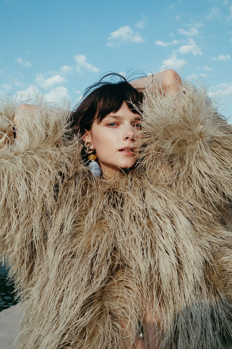 A woman in a beige feathered coat stands outdoors against a sunny blue sky with wispy clouds. She wears drop earrings and has bangs.