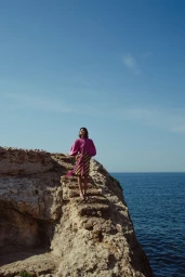 Model standing on a rocky coastal cliff wearing a bright pink blouse and striped skirt against a clear ocean view.