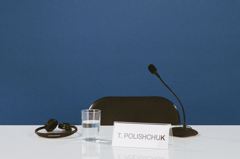 Meeting setup on a white table with a glass of water, microphone, headset, name card. Dark blue background. Conference scene.
