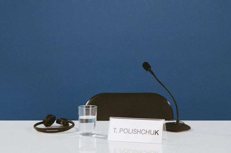 Meeting setup on a white table with a glass of water, microphone, headset, name card. Dark blue background. Conference scene.