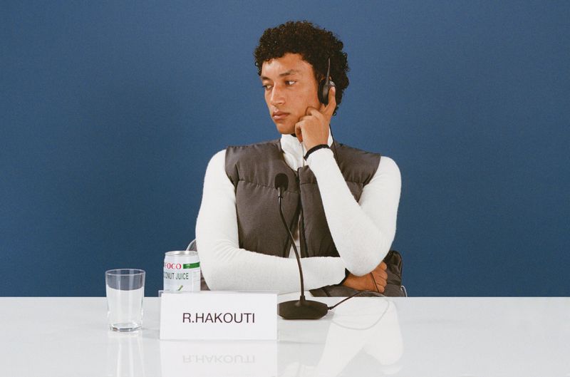 Man wearing headset sits at table with coconut juice, microphone in front of blue backdrop, looking away from camera.