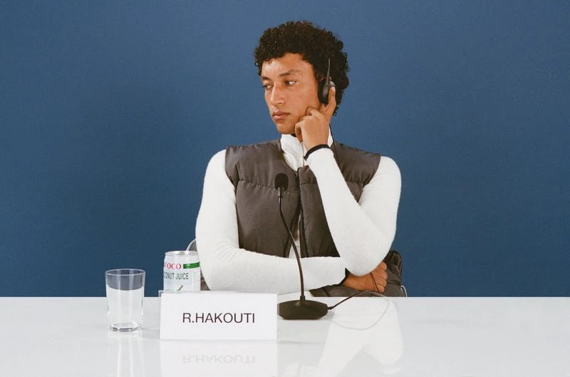 Man wearing headset sits at table with coconut juice, microphone in front of blue backdrop, looking away from camera. 