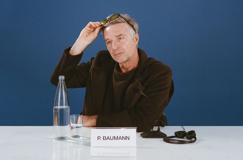 Adult male in a dark coat sits at a table with a water bottle, a glass, and headphones against a dark blue backdrop.