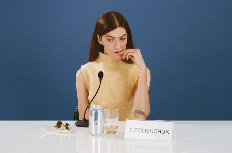 Woman in sleeveless top sits at a table with microphone, beverage can, and sunglasses against a solid blue background in a photo studio.