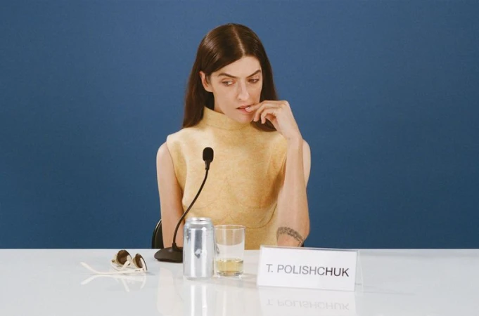 Woman in sleeveless top sits at a table with microphone, beverage can, and sunglasses against a solid blue background in a photo studio.