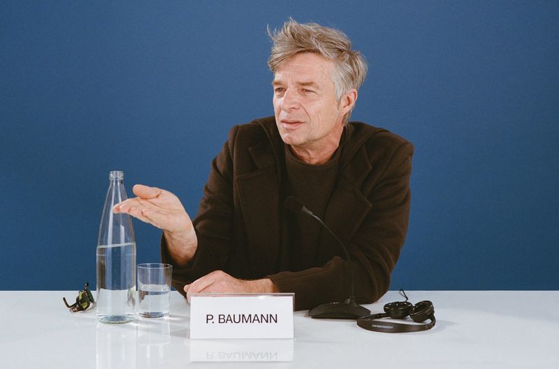 Mature man with grey hair and a dark blazer seated at a white table with a bottle of water, glass, microphone, and nameplate in studio.