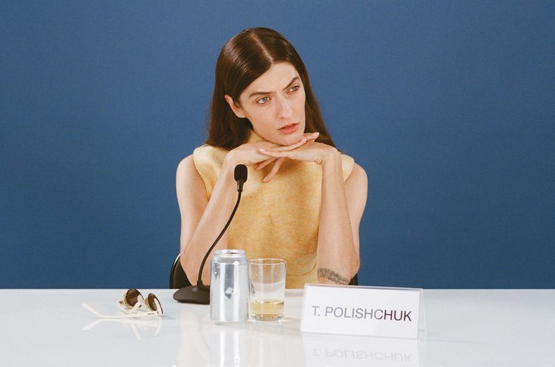 Singer, songwriter, and producer T. Polischuk sits at a desk with a can, sunglasses, and microphone against a solid blue backdrop.