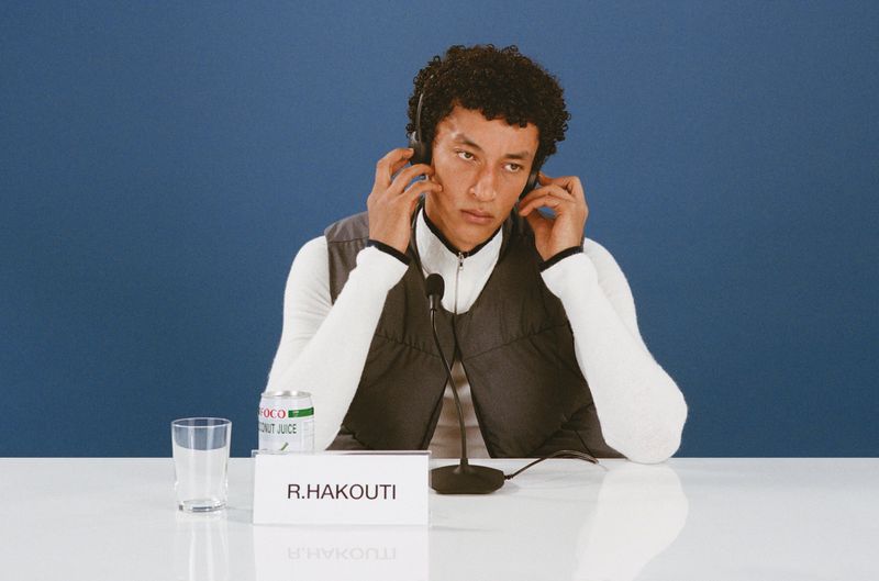 Adult male wearing headphones sits behind a white table with a microphone in front of a blue backdrop indoors in a medium shot.