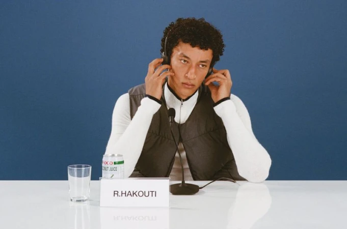 Adult male wearing headphones sits behind a white table with a microphone in front of a blue backdrop indoors in a medium shot.