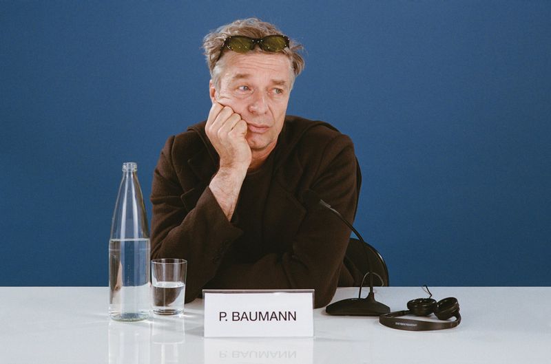 Man sits at a table with a bottle of water, a glass, and a microphone. He rests his face in his hand, lost in thought against a blue backdrop.