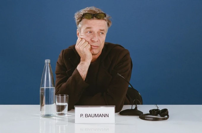 Man sits at a table with a bottle of water, a glass, and a microphone. He rests his face in his hand, lost in thought against a blue backdrop.