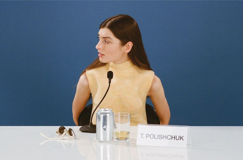 Portrait of a woman with long brown hair sitting at a white table in front of a microphone with a blue background, looking to the side.