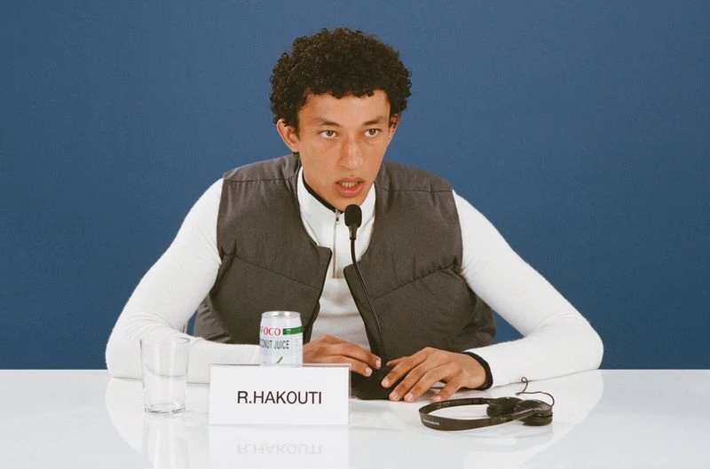 Adult male seated at a press conference table with microphone, glass, can of coconut juice, and headphones against a blue background.