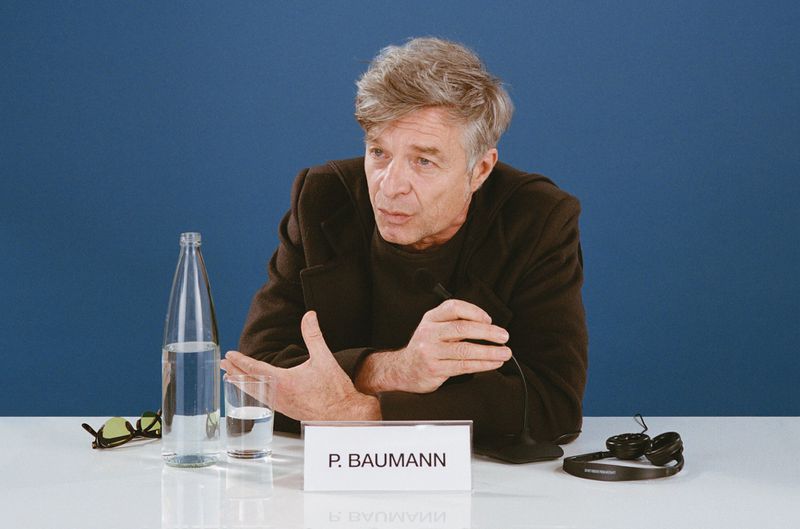 Mature adult man in dark jacket speaking at a table with microphone, water bottle, and glasses, against a blue background.