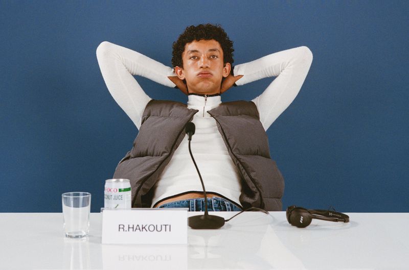 Adult male poses with hands behind his head wearing a gray vest, white turtleneck, and blue jeans sitting at a table against a blue background.