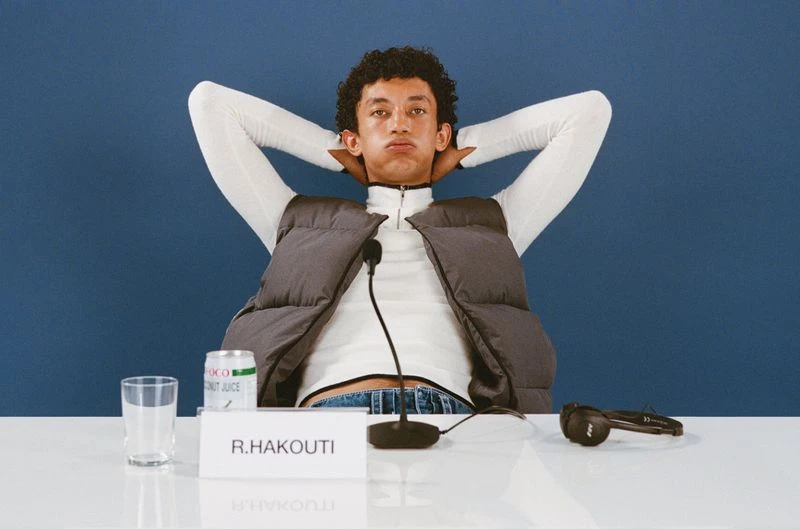 Adult male poses with hands behind his head wearing a gray vest, white turtleneck, and blue jeans sitting at a table against a blue background.