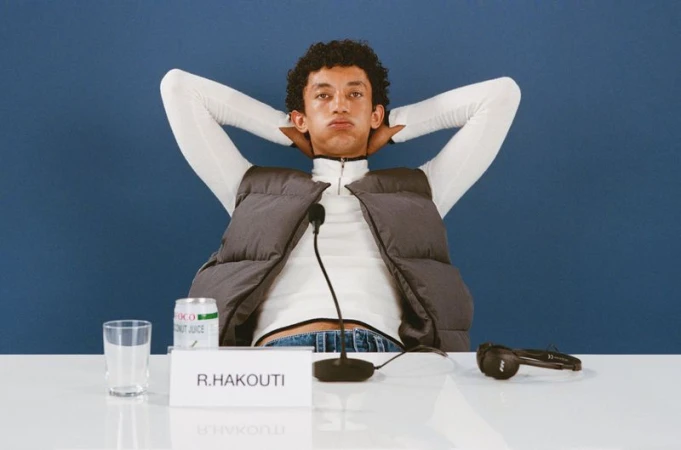 Adult male poses with hands behind his head wearing a gray vest, white turtleneck, and blue jeans sitting at a table against a blue background.