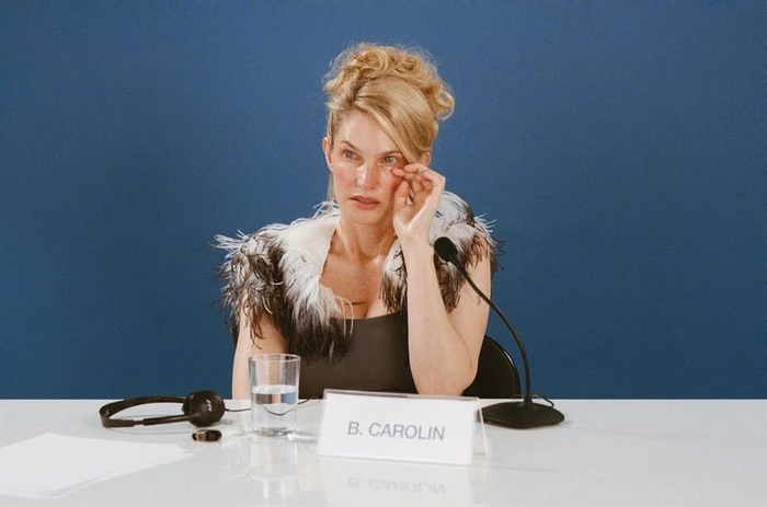 Woman in feathered collar sitting at a press conference table with microphone and nameplate reading B. Carolin