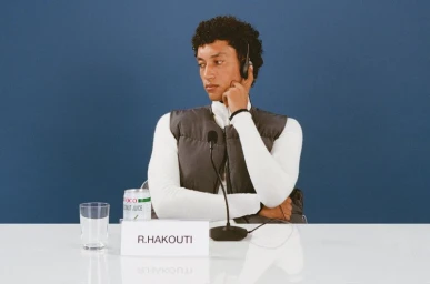 Person wearing a grey puffer vest and white top seated at a press conference table with a microphone against a blue backdrop.