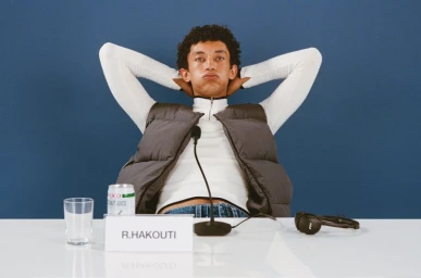 Young man with curly hair wearing a white turtleneck and dark puffer vest seated at a white desk with hands behind his head.