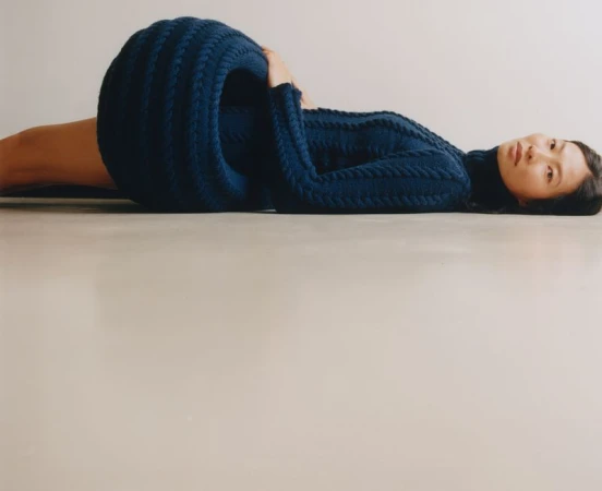 Model lying on a pale floor wearing a voluminous dark blue ribbed knit garment for Constance Tsang project by Present Space.