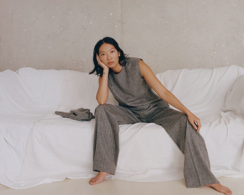 Adult woman poses in gray sleeveless top and matching pants on a white couch against a light gray wall in a studio setting.
