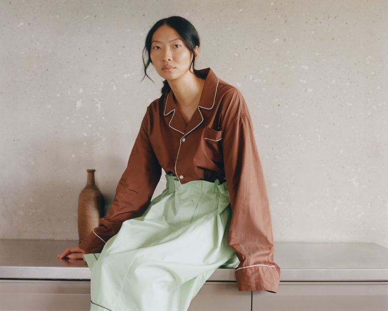 Portrait of a young Asian woman in a brown pajama top and light green skirt, sitting on a metal surface in front of a neutral backdrop.