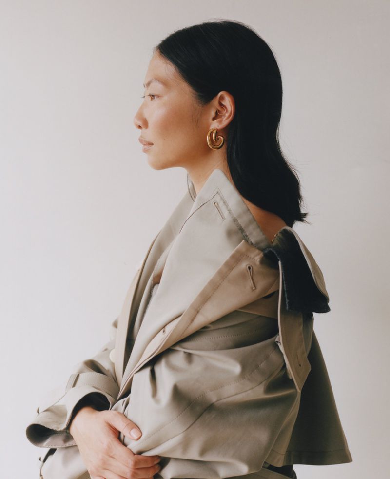 Elegant Asian woman in a tan trench coat and gold earrings, side view, with a clean off-white backdrop in a fashion or beauty setting.