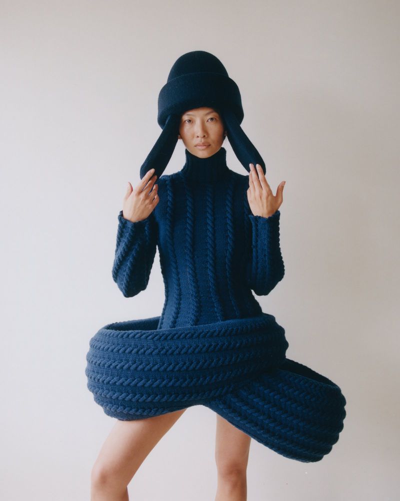 Asian fashion model in an oversized blue knitwear dress and hat poses in a photo studio against a neutral background for a fashion editorial.