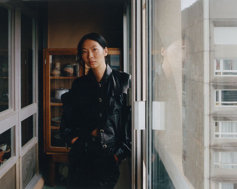 Portrait of an Asian woman in a black leather jacket indoors near a window, with a neutral expression and an apartment building outside.