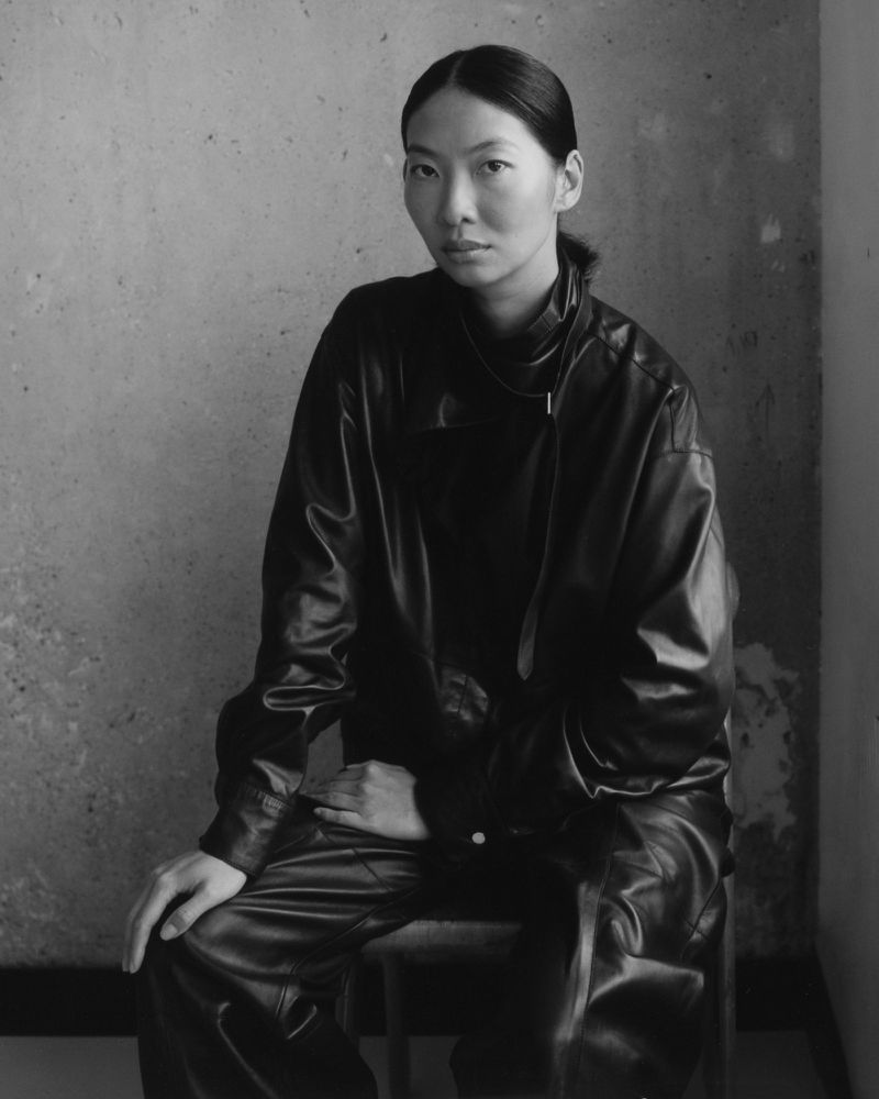 Portrait of an Asian woman in black leather jacket and pants sitting in front of a textured wall, illuminated by natural light, in monochrome.
