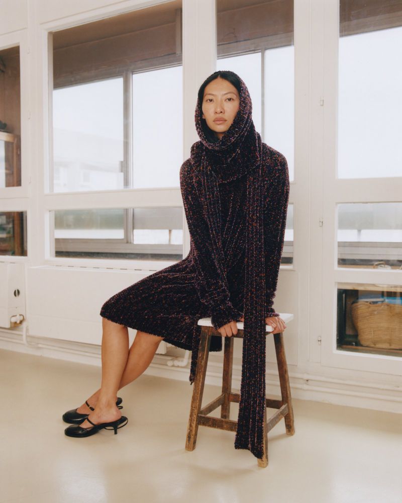 An Asian woman sits on a wooden stool in front of a window wearing a knit dress with a matching scarf draped over her head.