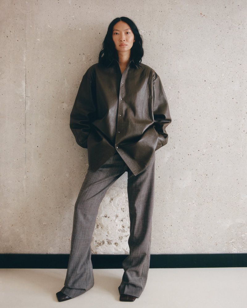 Adult woman in relaxed pose wearing an unbuttoned brown leather top and gray trousers standing in front of a concrete wall.
