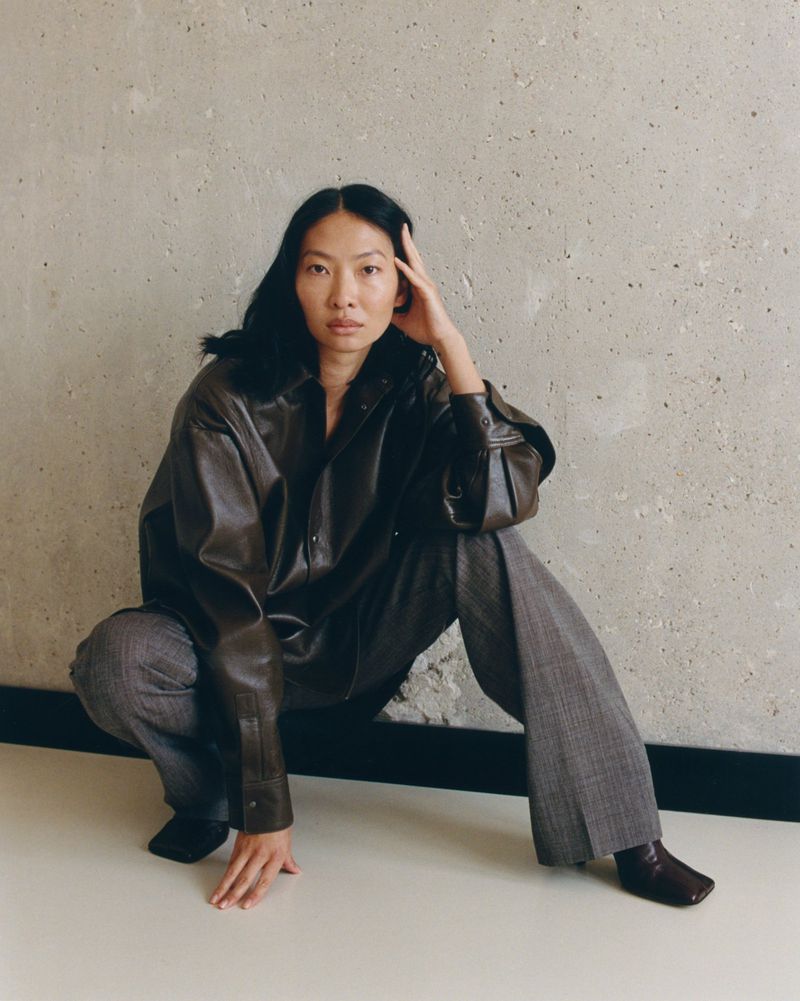 Asian woman in a brown leather jacket and plaid pants is crouching in front of a grey wall. Dark hair, neutral facial expression.