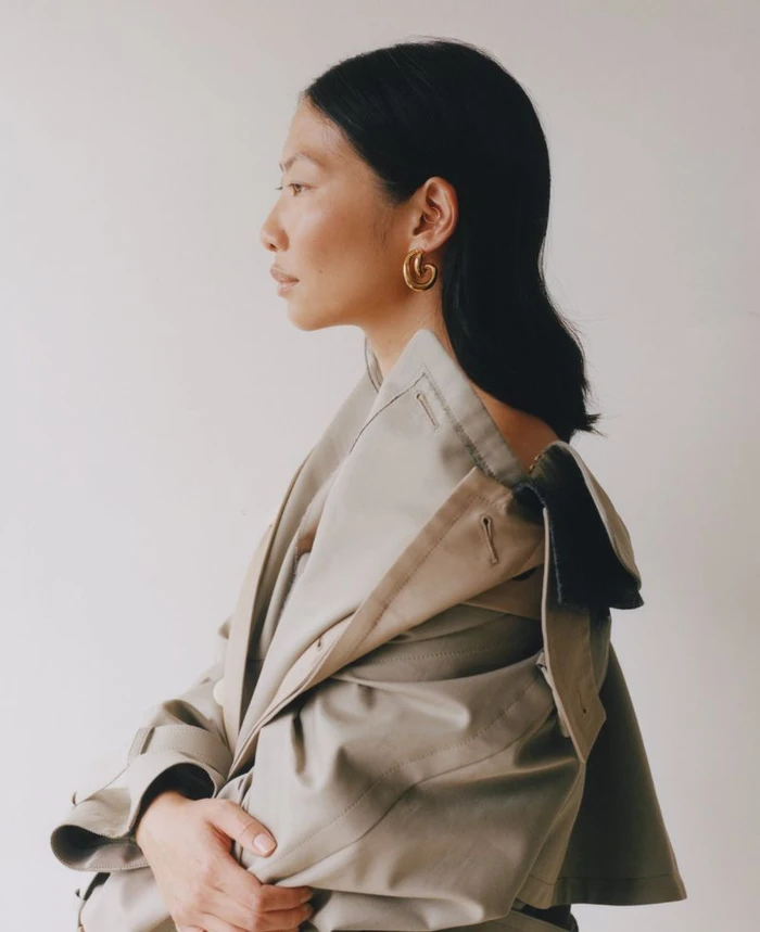 Profile portrait of a woman wearing a beige trench coat and gold hoop earrings against a neutral background.