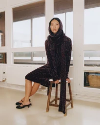 Model seated on a wooden stool wearing a dark knit dress with a matching hood and scarf, set against a multi-pane window.