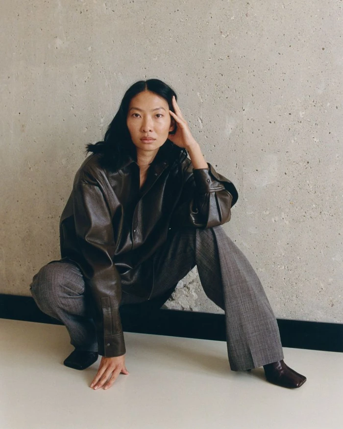 Woman crouching against a concrete wall wearing a dark brown leather shirt and grey plaid trousers