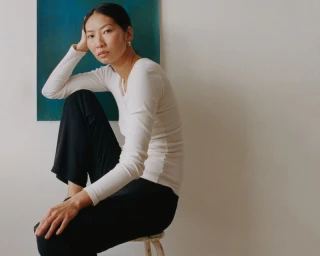 Asian woman seated on a small stool wearing a white long-sleeve top and black trousers against a plain wall with blue artwork