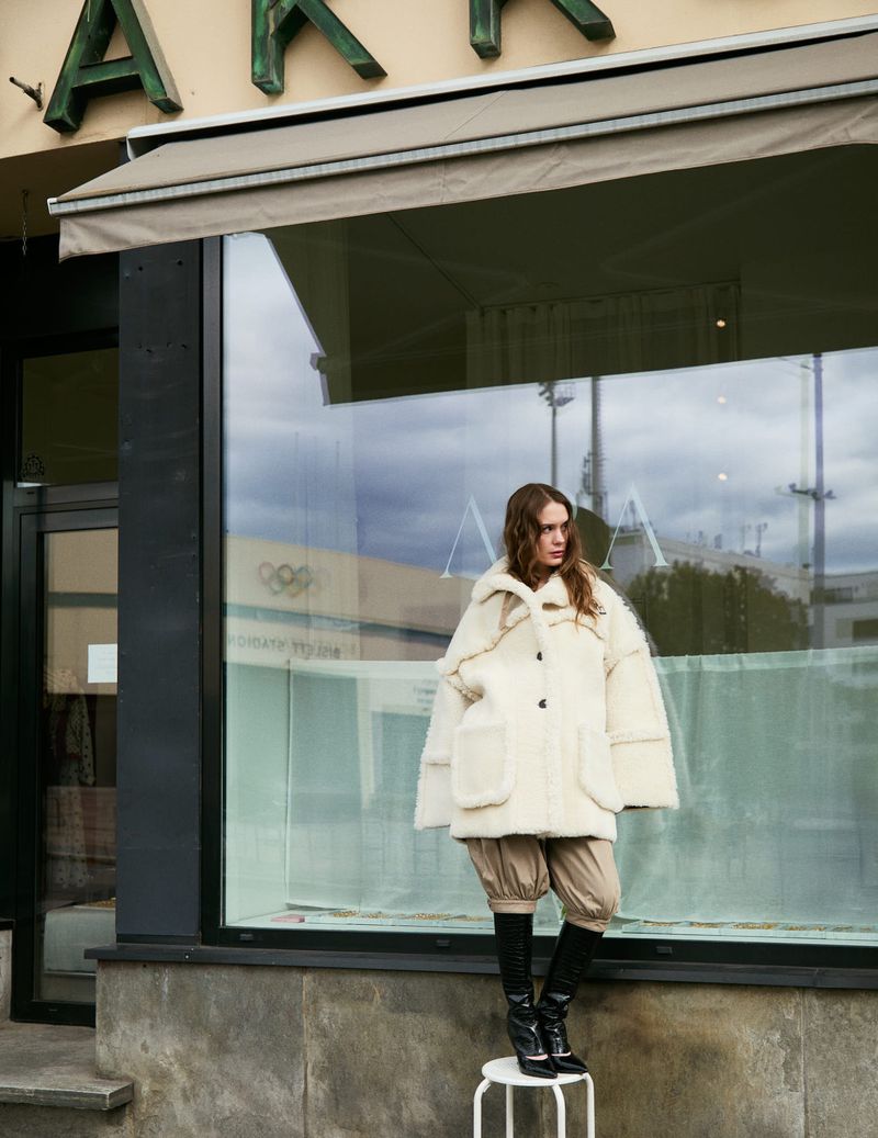 Fashion shot of a woman in an off-white coat and black boots posing on a stool outside a storefront with architectural details.