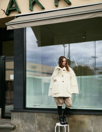 Fashion shot of a woman in an off-white coat and black boots posing on a stool outside a storefront with architectural details.