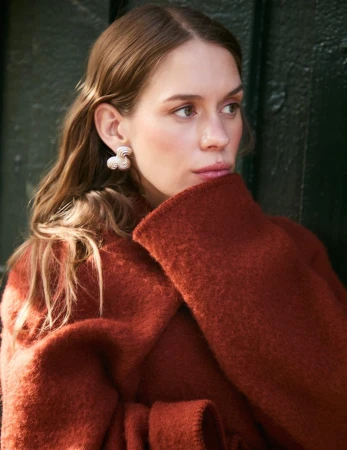 Portrait of a woman with long brown hair wearing modern silver earrings and a rust-colored wool coat with a blurred green background.