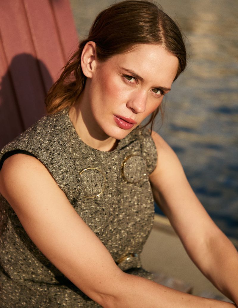 Portrait of an adult female in a tweed dress and minimal makeup, sitting outside near the water in neutral light.