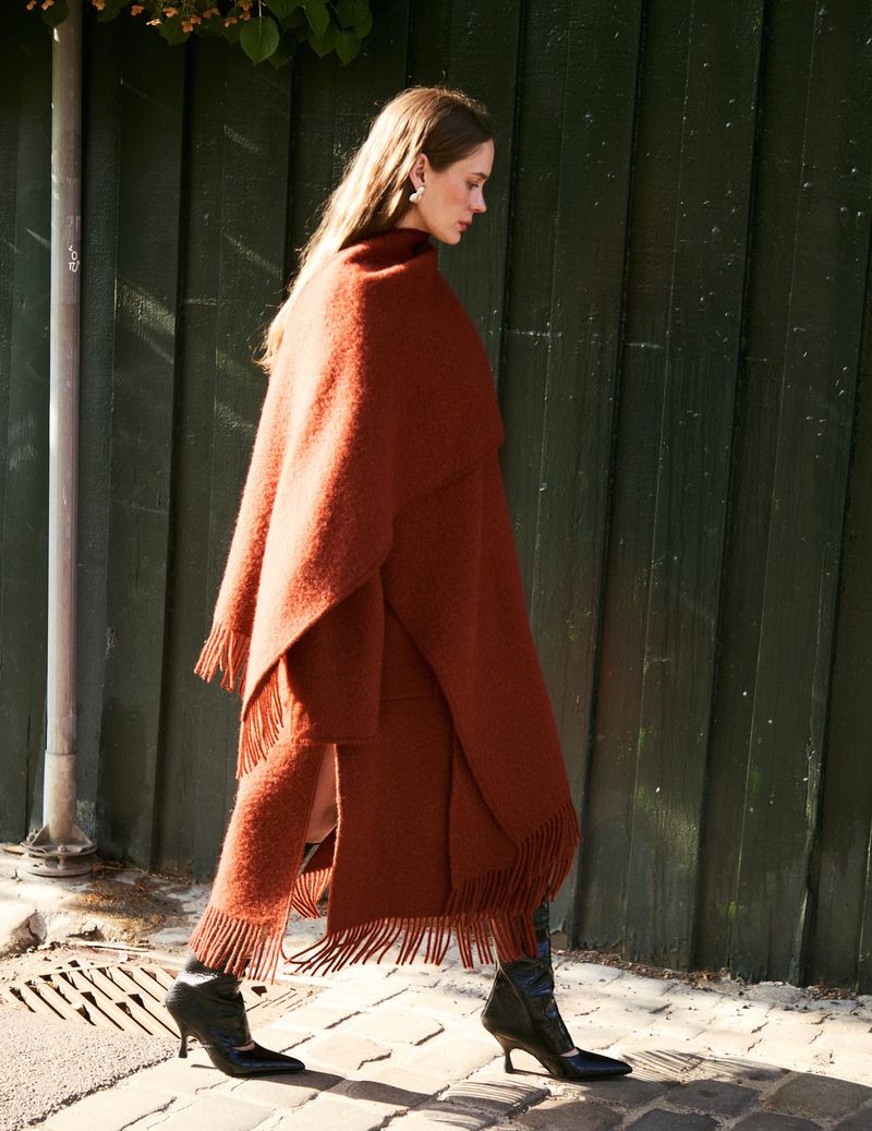 A woman walks alongside a green wall in a matching brown fringe-trimmed poncho and pants, paired with black heeled boots in natural light.