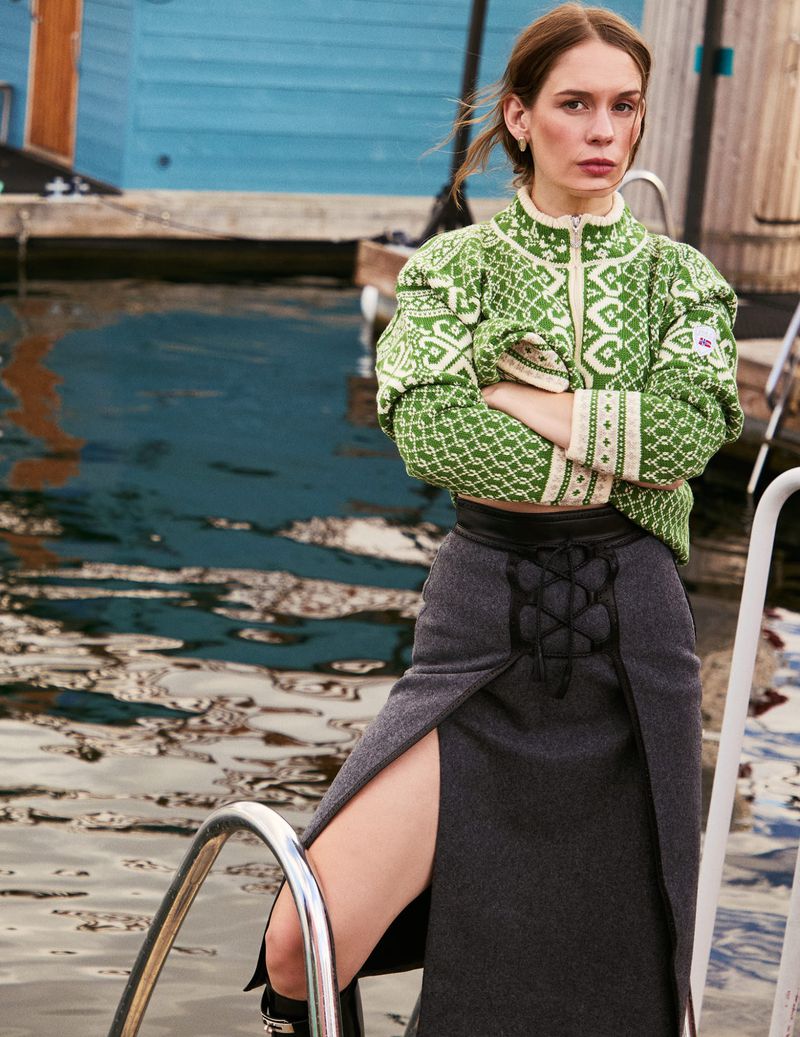 A woman with her arms crossed, wearing a green patterned jacket and grey skirt, standing on dock steps near water and a house.