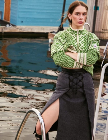 A woman with her arms crossed, wearing a green patterned jacket and grey skirt, standing on dock steps near water and a house.